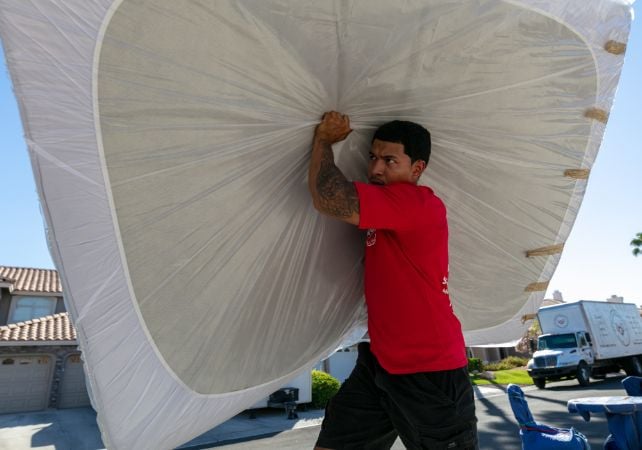 Man carrying large wrapped mattress outdoor.